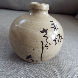 May include: A small, round, beige ceramic vase with black calligraphy writing on the side. The writing includes the number "5" and the characters "3, th".