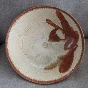 May include: A round ceramic bowl with a cream-colored base and a wide, textured rim in a deep reddish-brown hue. The interior features a radial pattern with a prominent, brush-like reddish-brown accent extending from the center to the edge.