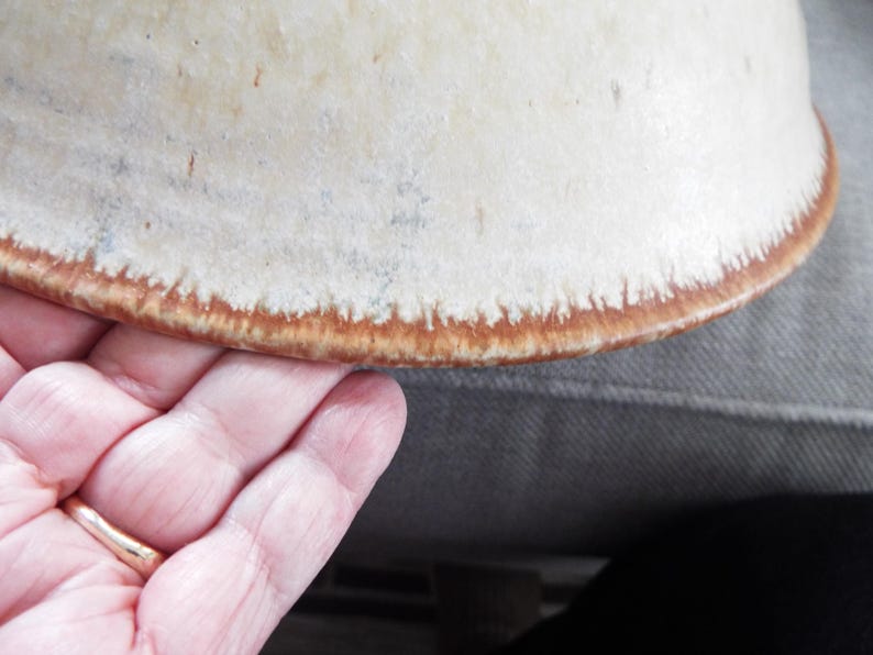 May include: A ceramic bowl with a cream-colored, speckled surface and a distinct, textured brown rim. The glaze transitions from a smooth, light off-white center to a darker, earthy brown edge with visible vertical drip-like patterns along the border.