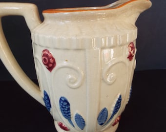 Puede incluir: Una jarra de cerámica de color crema con un borde marrón y diseños florales azules y rojos.