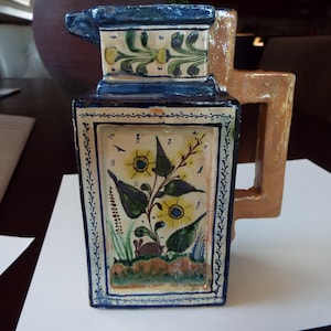 May include: A rectangular ceramic pitcher with a tan handle and a blue rim. The front panel features a floral design with yellow flowers, green leaves, and a small rabbit. The pitcher is decorated with blue and white patterns.