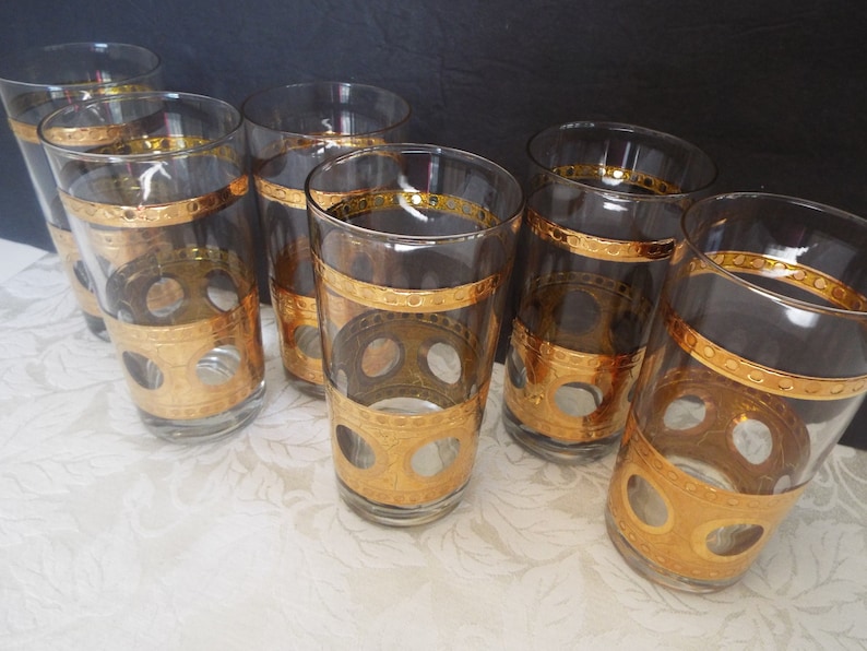 May include: Seven clear glass tumblers with gold trim and a pattern of circles. The glasses are arranged in a row on a white surface.