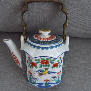 May include: A white porcelain teapot with a brass handle and a floral design. The teapot has a lid with a geometric pattern and a bird with a long tail.