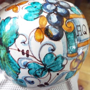 May include: A round ceramic vessel with a white base featuring hand-painted designs of teal leaves, dark blue grapes, and yellow accents. The surface displays intricate swirling patterns and the letters AQ in black ink, showcasing a glossy finish.