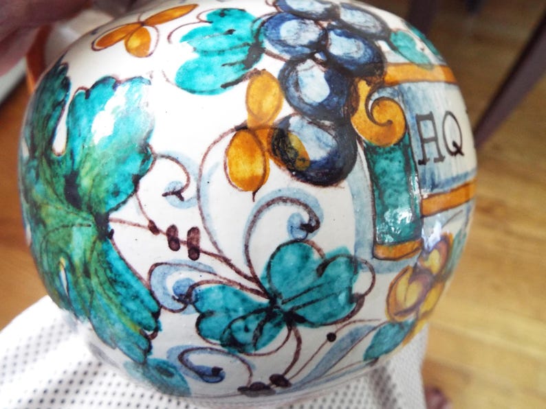 May include: A round ceramic vessel with a white base featuring hand-painted designs of teal leaves, dark blue grapes, and yellow accents. The surface displays intricate swirling patterns and the letters AQ in black ink, showcasing a glossy finish.