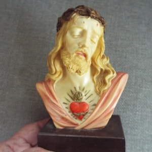 May include: A bust of Jesus Christ with a crown of thorns and a red heart on his chest. The bust is made of white and pink painted plaster and is on a wooden base.
