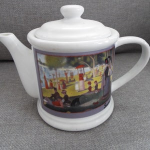 May include: White ceramic teapot with a lid and a handle. The teapot has a colorful image of people in a park on the side. The image includes trees, a pond, and a red umbrella. The text "Seurat" is visible at the bottom of the image.