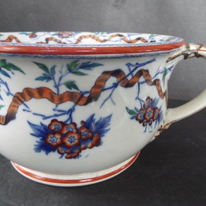 May include: A white ceramic chamber pot with a blue and orange floral design and a brown ribbon. The pot has a handle and a red rim.
