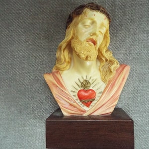 May include: A bust of Jesus Christ with a crown of thorns and a red heart with thorns on his chest. The bust is made of a light-colored material and is on a dark wood base.