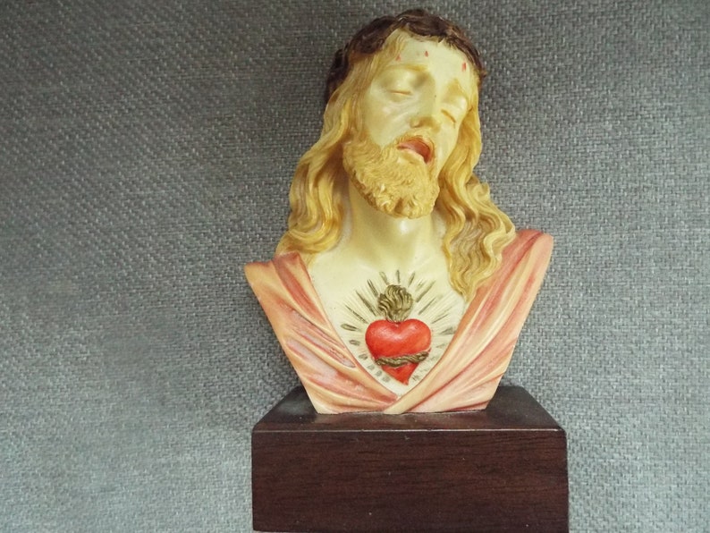 May include: A bust of Jesus Christ with a crown of thorns and a red heart with thorns on his chest. The bust is made of a light-colored material and is on a dark wood base.