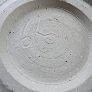 May include: The underside of a light gray ceramic vessel featuring a circular foot ring. A hand-carved maker's mark is impressed into the center of the base, consisting of a stylized spiral design with an elongated line extending from the center.