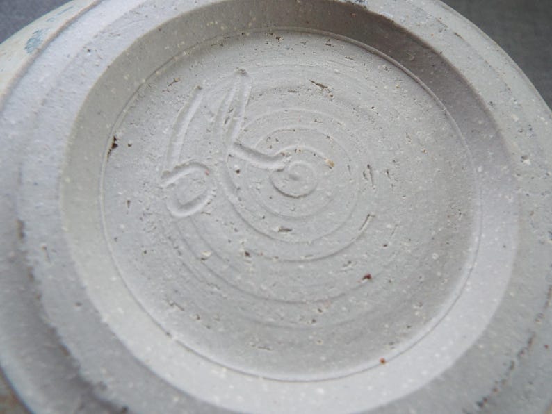 May include: The underside of a light gray ceramic vessel featuring a circular foot ring. A hand-carved maker's mark is impressed into the center of the base, consisting of a stylized spiral design with an elongated line extending from the center.