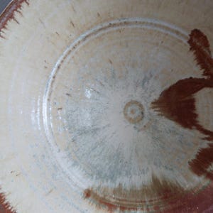 May include: A round ceramic bowl featuring a cream-colored base with a speckled texture. The rim and interior display a reddish-brown glaze that radiates toward the center, creating a feathered, brush-like pattern across the surface of the bowl.