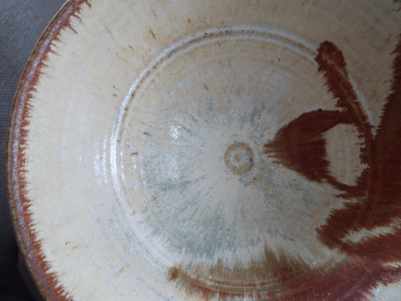 May include: A round ceramic bowl featuring a cream-colored base with a speckled texture. The rim and interior display a reddish-brown glaze that radiates toward the center, creating a feathered, brush-like pattern across the surface of the bowl.