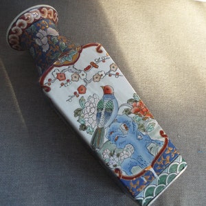 May include: A white porcelain vase with a square body and a rounded top. The vase is decorated with a colorful floral and bird design. The design features a blue bird with red accents, pink and white flowers, and green leaves. The vase has a gold trim around the top and bottom.