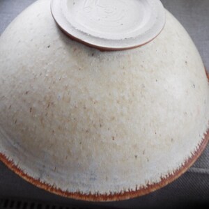 May include: A ceramic bowl shown from the underside, featuring a speckled off-white glaze with a textured surface. The circular base is raised and unglazed, while the outer rim displays a distinct reddish-brown band with a feathered edge.
