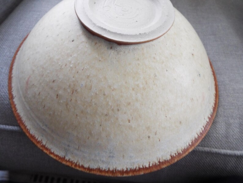 May include: A ceramic bowl shown from the underside, featuring a speckled off-white glaze with a textured surface. The circular base is raised and unglazed, while the outer rim displays a distinct reddish-brown band with a feathered edge.
