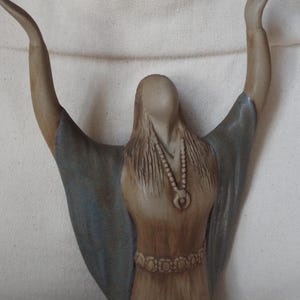 May include: A ceramic sculpture of a woman with her arms raised, wearing a long, flowing robe with blue and brown accents. She has a beaded necklace and a decorative belt.