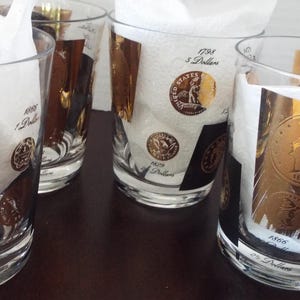 May include: Four clear glass tumblers with gold and black designs featuring images of coins. The glasses have the text "1798 5 Dollars" and "1800 5 Dollars" printed on them.