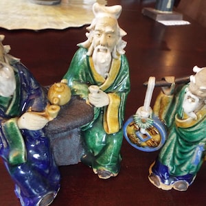May include: Three ceramic figurines of men with long white beards, wearing green, blue, and yellow robes. The figurines are sitting at a table with a teapot and cups.