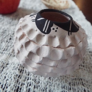 May include: A small, round, white ceramic pot with a black and white geometric design. The pot has a textured surface and a wide opening.