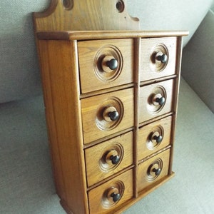 May include: A wooden spice rack with eight drawers, each with a black knob. The drawers are arranged in two columns of four. The rack is a light brown color and has a decorative top.