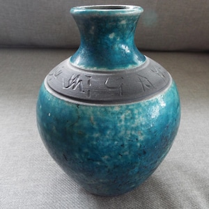 May include: A teal blue ceramic vase with a dark gray band around the middle. The band has an inscription in a dark gray color.