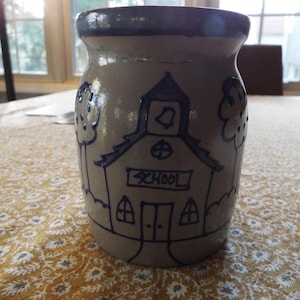 May include: A beige ceramic jar with a dark blue rim and a hand-drawn schoolhouse design. The schoolhouse has a bell tower, windows, and the word "SCHOOL" written across the front. Two trees flank the schoolhouse.