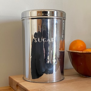 Puede incluir: Un recipiente de azúcar de metal plateado con la palabra "SUGAR" en relieve en la parte delantera. El recipiente está sobre una tabla de cortar de madera.