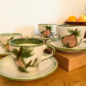 May include: A set of four ceramic teacups and saucers. The cups and saucers are off-white with a green rim and handle. Each cup features a hand-painted fig design in shades of pink, green, and brown. The set is arranged on a wooden surface.