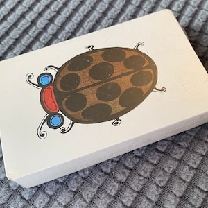 May include: A stack of white cards featuring a cartoon ladybug illustration. The ladybug has a brown body with black spots, a red head, and blue accents. The cards are stacked on a textured gray surface.