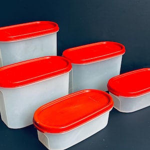May include: Five plastic food storage containers with red lids. The containers are oval-shaped and vary in size. The lids are a bright red, contrasting with the translucent white containers. The containers are arranged on a dark surface.