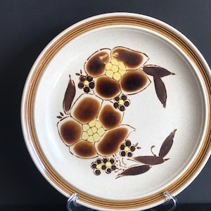 May include: A white ceramic dinner plate with a brown and yellow floral design. The plate has a brown rim with a raised edge.