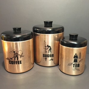 Puede incluir: Conjunto de tres botes de color cobre con tapas negras e ilustraciones en blanco y negro. Los botes están etiquetados como "Café", "Azúcar" y "Té".