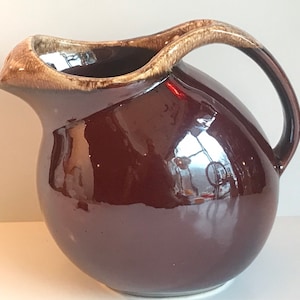May include: A brown ceramic pitcher with a wide, round body and a handle. The pitcher has a brown glaze with a light brown rim.