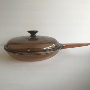 May include: A brown glass frying pan with a lid and a brown handle. The pan is made of tempered glass and is designed for cooking on the hob.