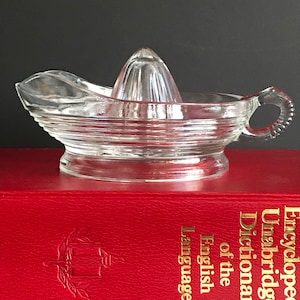 May include: A clear glass citrus juicer with a ribbed bowl, a pointed reamer, and a handle. The juicer sits on a red book with gold lettering. The background is dark.