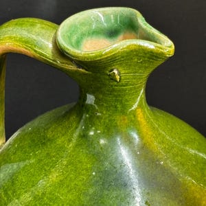 May include: A handcrafted, green ceramic pitcher with a curved handle and a wide, flared spout. The pitcher has a glossy finish with variations in the green glaze, showing hints of yellow and brown. The design is simple and elegant.