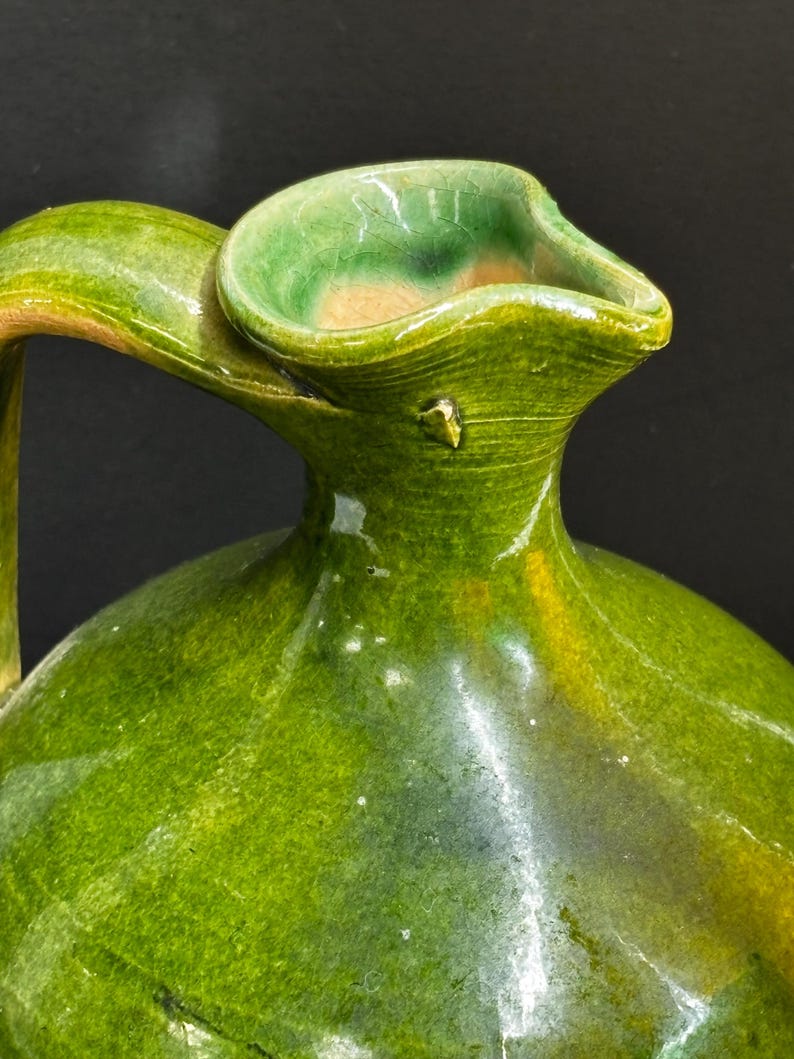 May include: A handcrafted, green ceramic pitcher with a curved handle and a wide, flared spout. The pitcher has a glossy finish with variations in the green glaze, showing hints of yellow and brown. The design is simple and elegant.