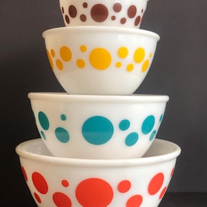Può includere: Set di quattro ciotole per mescolare bianche con pois di diversi colori. Le ciotole sono impilate in ordine di dimensione, con la ciotola più grande in basso e la ciotola più piccola in alto. I colori dei pois sono rosso, turchese, giallo e marrone.