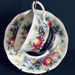 以下が含まれることがあります： ヴィンテージのティーカップとソーサーのセット。白い磁器のカップとソーサーには、赤いリンゴ、紫色のブドウ、青いバラを使ったカラフルなフルーツと花の模様が施されています。カップの内側は黒で、金色のトリムが付いています。