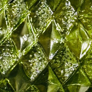 May include: Close-up of a textured, green glass surface. The surface is covered in a repeating diamond pattern, with each diamond having a raised, textured appearance. The glass reflects light, creating highlights and shadows.