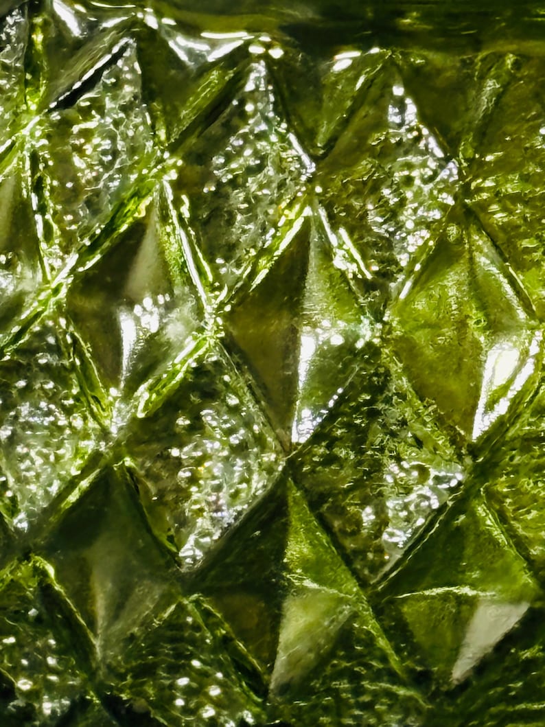 May include: Close-up of a textured, green glass surface. The surface is covered in a repeating diamond pattern, with each diamond having a raised, textured appearance. The glass reflects light, creating highlights and shadows.