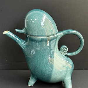 Puede incluir: Una tetera de cerámica turquesa con un diseño único y redondeado. La tetera presenta un asa curva con un detalle en espiral, un pico y una tapa. Se apoya sobre tres pequeños pies y tiene un acabado brillante, ideal para servir té.