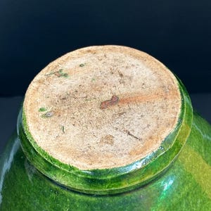May include: A close-up of the bottom of a green glazed ceramic pot. The base is a light brown color with a rough texture. The green glaze has variations in color, creating a textured appearance.