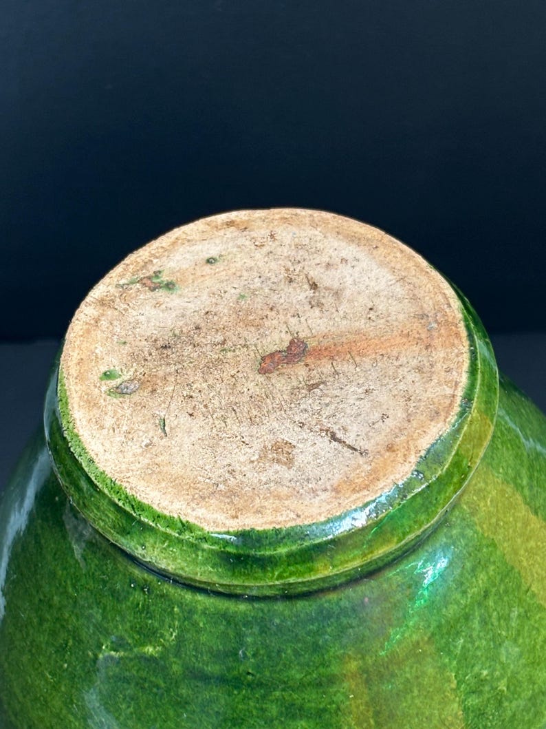 May include: A close-up of the bottom of a green glazed ceramic pot. The base is a light brown color with a rough texture. The green glaze has variations in color, creating a textured appearance.