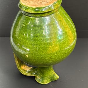 May include: A green ceramic jar with a rounded body, a small handle, and a lid. The jar has a glossy finish with variations in the green color. The lid is a light brown color. The jar is likely used for storage or decoration.