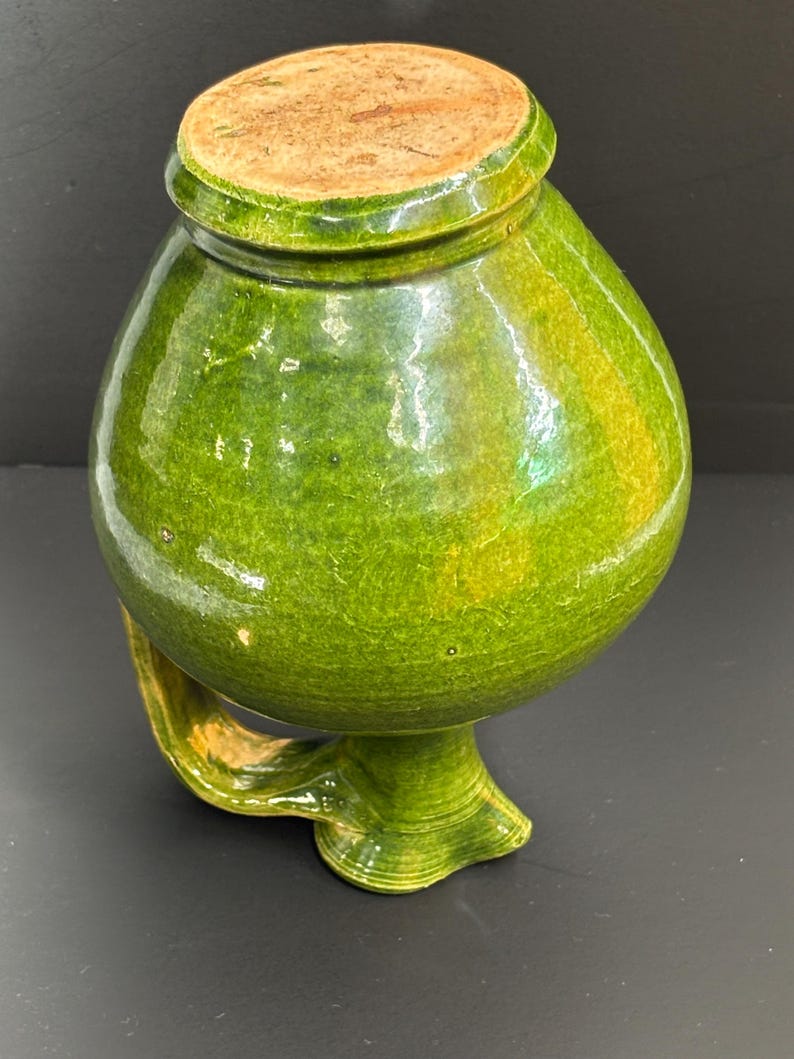 May include: A green ceramic jar with a rounded body, a small handle, and a lid. The jar has a glossy finish with variations in the green color. The lid is a light brown color. The jar is likely used for storage or decoration.