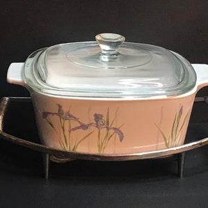 Pode incluir: Uma travessa quadrada branca com uma tampa de vidro e um desenho floral de íris roxas sobre um fundo castanho claro. A travessa está sobre um suporte de metal prateado com uma pega curva.