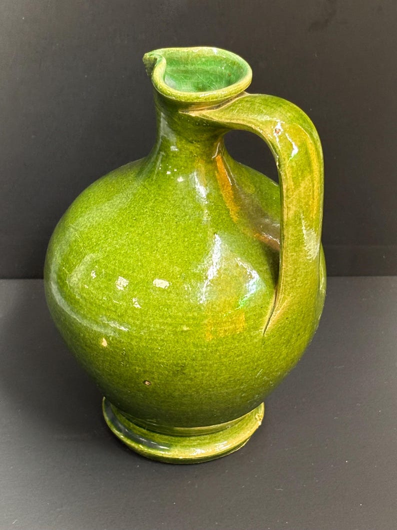 May include: A green ceramic pitcher with a rounded body, a curved handle, and a spout. The pitcher has a glossy finish and a base. The color is a vibrant, bright green.